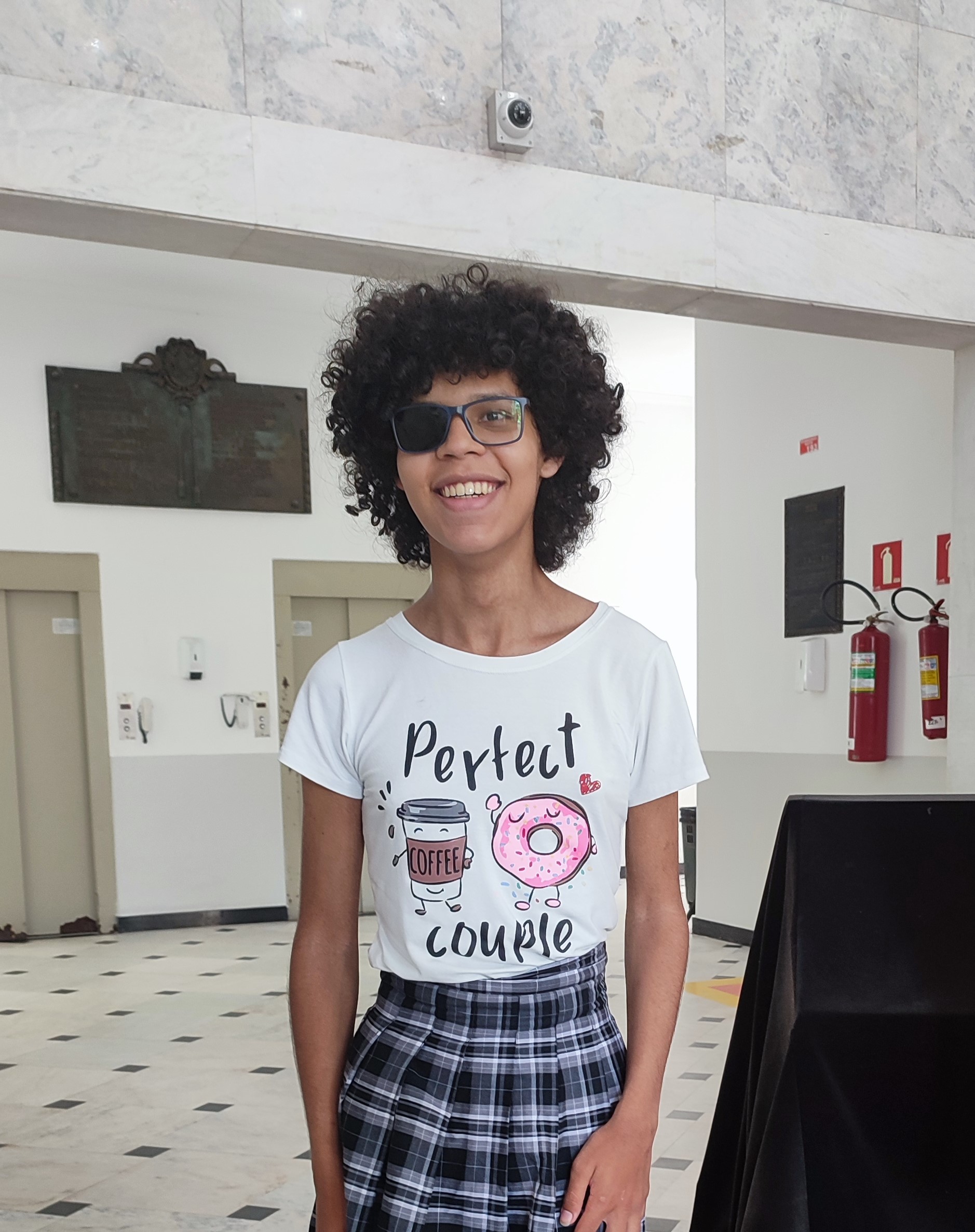 Foto da Larissa Ember, com uma camiseta branca escrita 'casal perfeito', saia cinza xadrez.